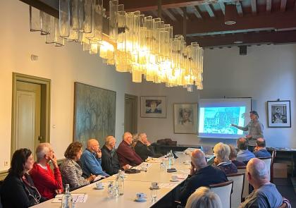 Frank geeft een lezing in een zaal in het stadhuis. Voor hem staat een lange tafel waar mensen aan zitten