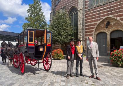 Links staat een 19e eeuwse koets geparkeerd voor de Grote- of Maria Magdalenakerk. Wethouder Martijn Vermeulen, archivaris Frank de Klerk en burgemeester Marcel Fränzel poseren voor de foto.